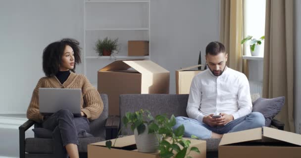 Εστιασμένη afro γυναίκα browses laptop σε νέο επίπεδο, καυκάσιος άνδρας καλεί μέσω smartphone, κινούνται σε νέο διαμέρισμα μαζί με τη γυναίκα, κάθονται σε άνετο καναπέ, που περιβάλλεται από κουτιά από χαρτόνι στο σαλόνι — Αρχείο Βίντεο