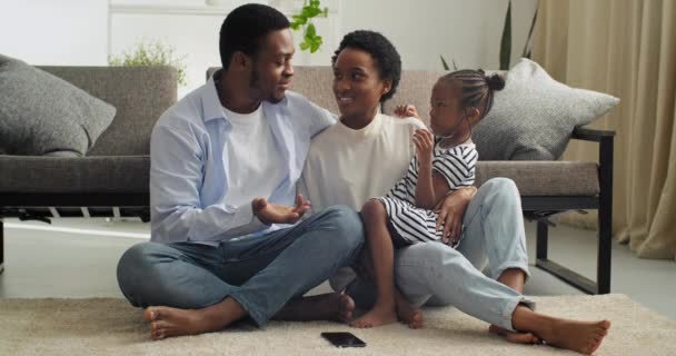 Üç çıplak ayaklı çift, bebek Afro-Amerikan aile babası ve tatlı küçük kız çocuğu yumuşak sıcak halıda oturmuş hafta sonları sohbet ediyorlar. — Stok video