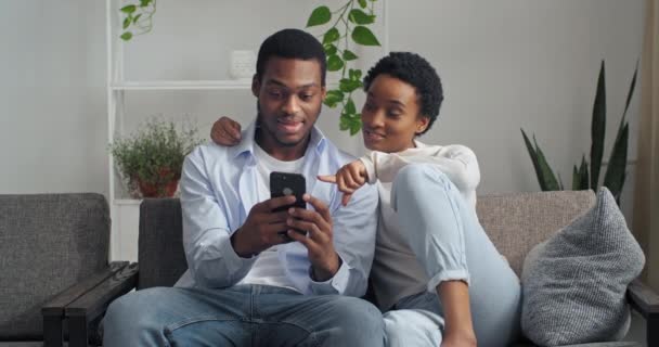 Afro amerykański mężczyzna i kobieta z krótkimi stylowe fryzury w modnym białym swetrze patrząc na telefon komórkowy urządzenie szuka informacji czytając złe wiadomości omawiając rozmowy siedzi w domu na przytulnej kanapie — Wideo stockowe