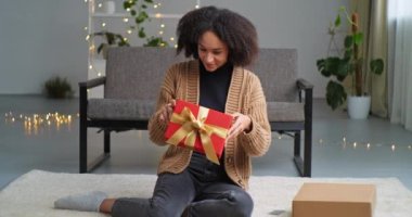 Genç gülümseyen Afro-Amerikan melez kadın oturma odasında oturmuş elinde kırmızı hediye kutusu olan kuryeyle hediye paketini alıyor.