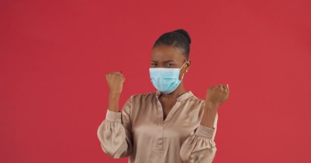 Портрет африканської успішної вродливої жінки, яка радіє закінченню карантину, носить захисну маску на обличчі від респіраторних захворювань, робить жест перемоги своїми руками, що святкує — стокове відео