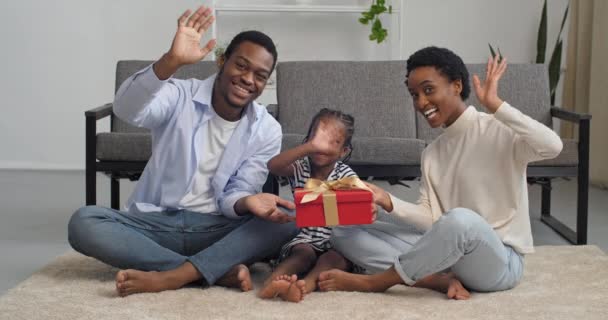 Οικογενειακό πορτραίτο στο σπίτι, τρία άτομα αφρικανικό ζευγάρι με παιδιά γονείς και μικρή χαριτωμένη κόρη με κόκκινο κουτί δώρου κάθεται ξυπόλητη στο πάτωμα στο χαλί κουνώντας χαιρετισμό χέρια κοιτάζοντας κάμερα χαμογελώντας — Αρχείο Βίντεο