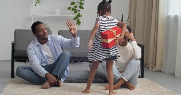 Famille afro-américaine, petite fille mignonne en robe se tient avec son dos à la caméra tenant boîte cadeau rouge dans ses mains regarde mère heureuse qui a fermé ses yeux donne cinq à papa assis sur le sol — Video