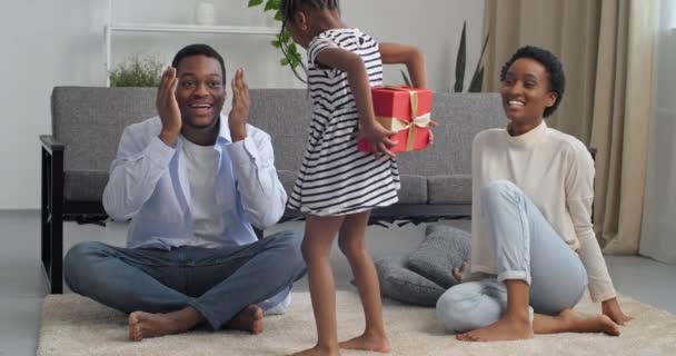 Trois personnes pieds nus famille à la maison dans le salon intérieur, petite fille afro-américaine généreuse avec fille tenant boîte-cadeau rouge derrière le dos et tenant à papa fait la surprise pour la fête des pères — Video