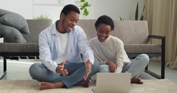 Glückliches afroamerikanisches Paar schaut auf Laptop beim Online-Kauf und spricht auf Videokonferenz gute Nachrichten lesen positive meil Freude Lotterie spielen, Mann und Frau sitzen auf dem Boden — Stockvideo