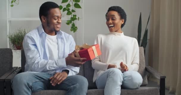 Mooi jong Afro-Amerikaans echtpaar dat thuis op de bank zit. Knappe zwarte man guy geven zijn vriendin atractive vrouw etnische dame rood cadeau doos cadeau feliciteert gelukkige verjaardag — Stockvideo