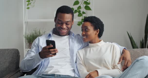 Mutlu genç koca, sosyal ağda cep telefonuyla komik videolar gösteriyor güzel eş. Gülümseyen Afro-Amerikan eşleri internetten alışveriş yapan, evdeki kanepede dinlenen çiftler. — Stok video