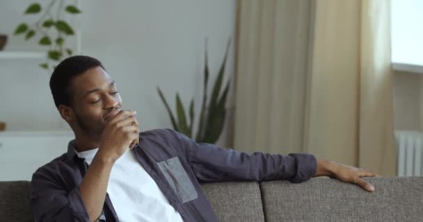 Portretul tânărului afro-american de rasă mixtă bărbat purtând haine de cămașă casual așezat pe o canapea confortabilă de mobilier modern odihnindu-se în camera de zi a casei căutând pensiv pe fereastră privind pe cineva — Videoclip de stoc