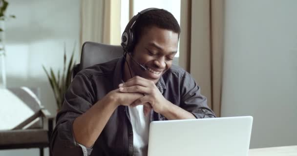 Крупным планом счастливый мужчина афро-американец молодой парень носит наушники и головной микрофон использует ноутбук, чтобы сделать онлайн видео-чат конференции онлайн во время коронавируса улыбается смеясь разговаривая — стоковое видео
