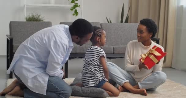 Padres afroamericanos cuidando a mamá y padre cariñoso hablando con su hija pequeña dándole regalo de cumpleaños a la caja de regalo roja para niños preguntándole a la chica sobre su comportamiento, familia celebrando sentada en el piso de la sala de estar — Vídeo de stock