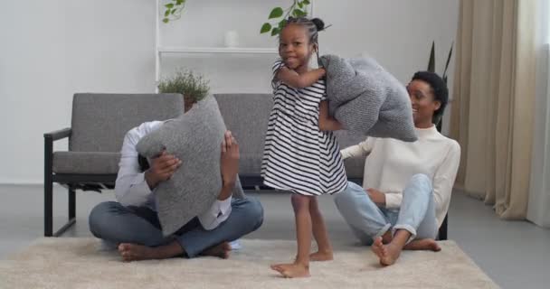 Joyeux jeunes parents afro-américains de la famille avec une petite fille enfant se sentent ludique engagés dans une bataille d'oreillers dans le salon, père et mère avec un petit enfant d'âge préscolaire impliqué dans le jeu sur le sol — Video