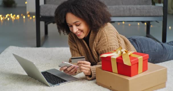Afro femme américaine consommateur se trouve sur le sol sur le ventre en face de l'ordinateur portable près boîte cadeau rouge achète des cadeaux en ligne en utilisant l'annuaire de téléphone intelligent mobile dans les billets de magasin en ligne détient la carte de crédit bancaire dans sa main — Video