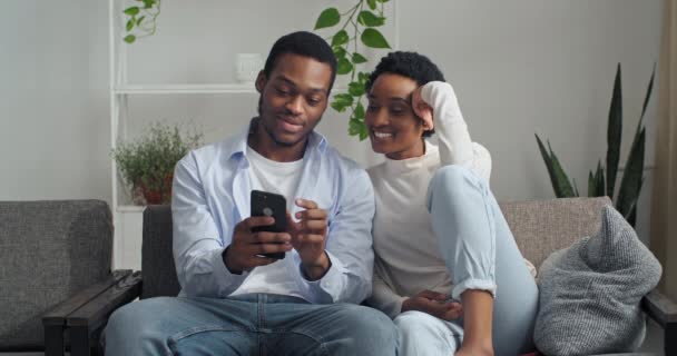 Afro-americano casal homem e mulher sentados juntos em casa olhando para a tela do telefone usando dispositivo móvel gadget para procurar bilhetes de reserva para férias escolhendo hotel na internet discutindo falar — Vídeo de Stock