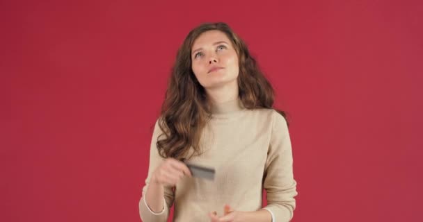 Студійний портрет молодої білої жінки красива дівчина в светрі, що показує пластикову банківську кредитну картку і дивиться на простір копіювання з урахуванням ідеї подарунка онлайн-замовлення ізольовано на червоному тлі — стокове відео