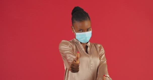 Retrato de sostenes afroamericanos de chica enmascarada. Alegre mujer lleva máscara médica en la cara muestra el pulgar hacia arriba, gesto como, símbolo de buena elección de pie aislado sobre la pared roja cansado de coronavirus — Vídeos de Stock