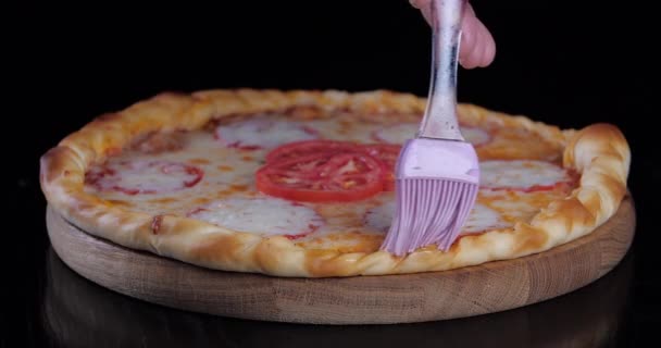 Close-up mano humana de grasas de chef con cepillo con bordes de mantequilla de queso de deliciosa pizza chatarra con queso y tomates prepara plato vegetariano de comida rápida acostado en la mesa sobre fondo negro oscuro — Vídeo de stock