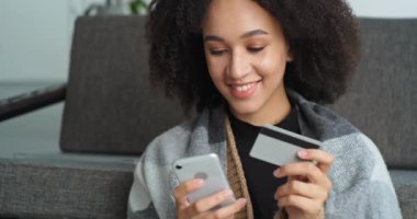 Portre gülümseyen Afro-Amerikalı kadın cep telefonuyla online alışveriş yapıyor akıllı telefon elinde ve kredi kartı eşya kitapları seyahat biletleri alıyor kuryeyle eve dönüyor.