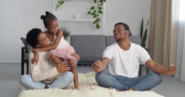 Афро-американская молодая женщина мама и черный мужчина папа сидеть босиком на полу гостиной в уютном доме медитировать маленькая милая этническая дочь активная девочка вмешиваться с родителями, делая упражнения йоги обнимает мать — стоковое видео