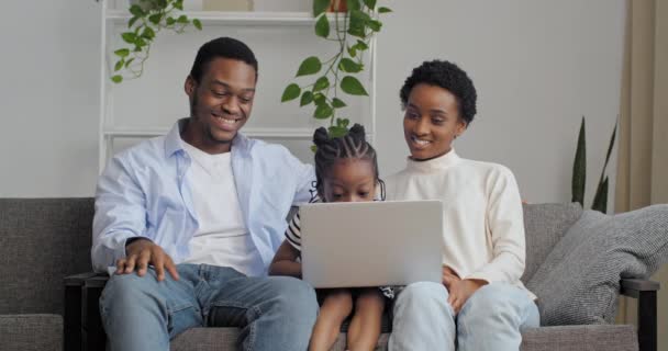 Home portrét mladých afrických amerických rodinných rodičů s malou dcerou sedí v obývacím pokoji interiér v útulném domě při pohledu na obrazovce notebooku sledování filmů karikatury videa on-line prostřednictvím počítače — Stock video