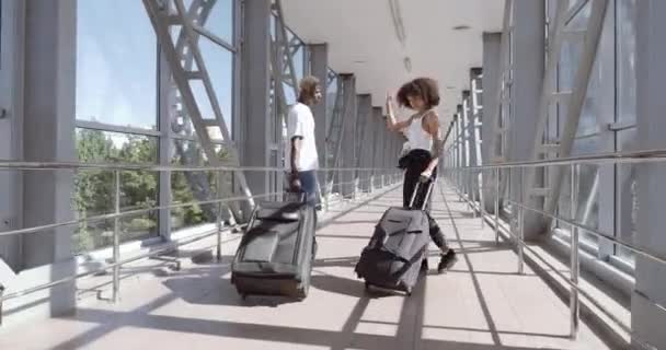 İki kişi abi ve kız kardeş arkadaş birkaç Afrikalı Amerikalı genç valiz taşıyorlar dans pisti boyunca havaalanı terminali koridorunda yürüyorlar uçakla eve dönerken sevinçle uçuyorlar. — Stok video