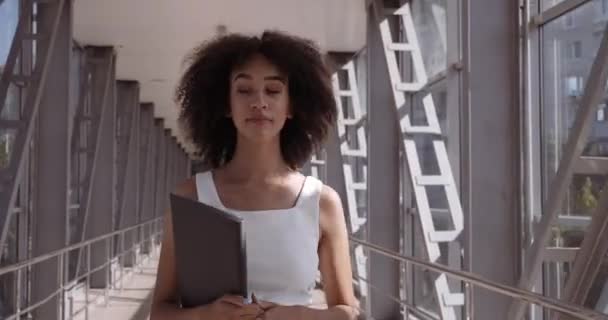 Αυτοπεποίθηση σκεπτική επιχειρηματίας διευθυντής φοιτητής γραμματέας αφροαμερικανός γυναίκα με σγουρά κομψό χτένισμα με μαύρο φάκελο με έγγραφα περπατά κατά μήκος μπαλκόνι του περάσματος στο γραφείο κατά μήκος του διαδρόμου — Αρχείο Βίντεο