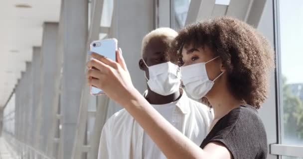 Casal afro-americano em máscaras de proteção médica dispara vídeo para blog vlog no telefone tirar foto on-line contra fundo da conversa da cidade por vídeo chat conferência de comunicação remota com amigos — Vídeo de Stock