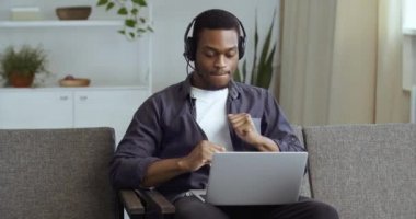Kulaklıklı, bilgisayar ekranında müzik dinleyen, evinde kanepede oturmuş dans eden Afro-Amerikan genç ve mutlu bir adamın portresi ellerini melodinin ritmine göre hareket ettiriyor.