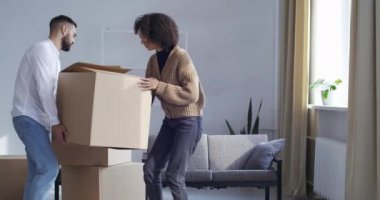 Çok ırklı çift kıvırcık saçlı afro kadın ve beyaz adam oturma odasına büyük bir karton kutu taşıyorlar, genç bir aile eşyaları yeni evlerine taşıyor, gayrimenkul teslimatı veya konsept satın alıyorlar.