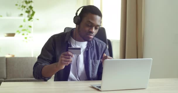 Zakenman maakt online kopen terwijl zittend achter laptop houdt bank credit card in zijn hand voert geheime code voor het bestellen op internet luisteren naar muziek op koptelefoon, e-commerce sale concept — Stockvideo