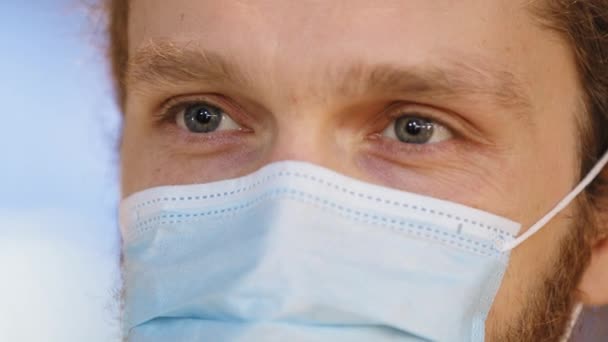 Primer plano de la cara masculina en máscara quirúrgica de protección médica. Retrato de hombre caucásico médico paciente con ojos azules lleva respirador de la infección con virus covid mirando a la cámara, concepto de peligro — Vídeos de Stock