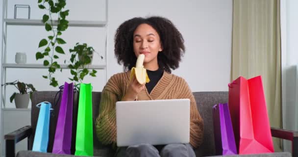 Afro-amerikan içerikli aç kadın kanepenin üzerinde oturmuş dizüstü bilgisayarlı paketler tutuyor sağlıklı muz meyveleri yiyor internetten mal sipariş ettikten sonra başparmağını doğrultuyor. — Stok video