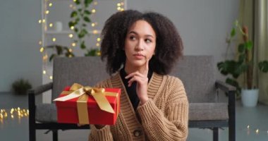 Düşünceli Afro-Amerikan kadın, şık kıvırcık saçlı, kırmızı hediye kutusuyla yerde oturuyor. Doğum günü hediyesi olarak. Paketin içinde ne olduğunu düşünüyor.
