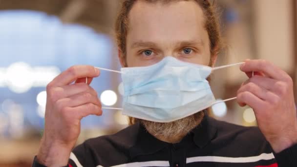 Porträtt av kaukasisk tusenårig man sätta på skyddande medicinsk mask på ansiktet blinkar med ögat, närbild kille bär respirator skyddar hälsan från att infekteras med covid coronavirus pandemi — Stockvideo