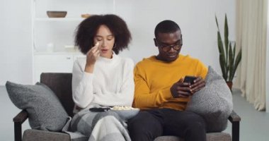 Afro-Amerikalı çift arkadaşlar oturma odasında oturmuş patlamış mısır yiyorlar radyoda müzik dinliyorlar dans ediyorlar, el ele tutuşup internette akıllı telefon tutuyorlar.