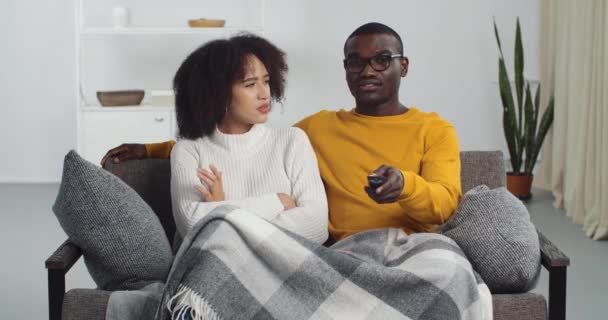 Młoda para kłócąca się oglądająca telewizję siedzącą na kanapie w domu salon, szczęśliwy Afrykanin mąż chłopak śmiejący się z programu komediowego patrzy na niezadowoloną zdenerwowaną żonę siedzącą z skrzyżowanymi ramionami — Wideo stockowe