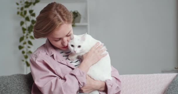 Молода біла дівчина з коротким волоссям власник кішки сидить на дивані зі своєю улюбленою твариною обіймає тварину ніжно приборкує концепцію любові і домашнього затишку, відпочиває на вихідних у вітальні або ввечері після роботи — стокове відео