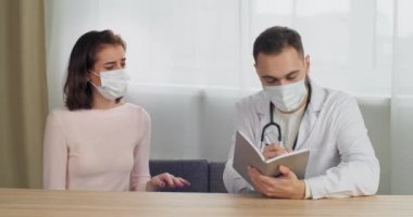 Beyaz erkek uzman doktor tıbbi maske takıyor defter muayenelerinde kadın hasta hakkında şikayetler yazıyor esmer kadın omuz ağrısı hakkında sorular soruyor, salgın sırasında klinikte buluşuyor.