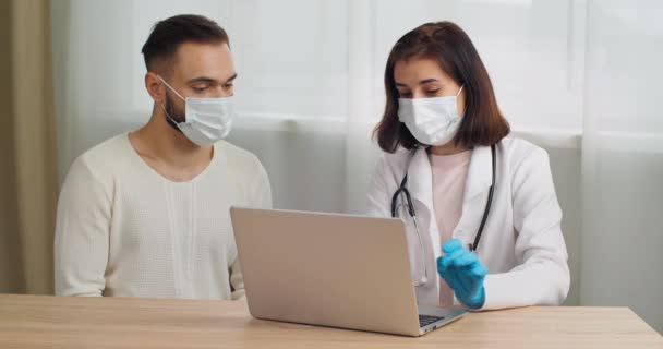 Mujer médico de bata blanca lleva guantes de látex mascarilla aconseja paciente, hombre quejándose de dolor de pecho, especialista escribiendo en la cita médica portátil explica, visita al terapeuta en el hospital clínico — Vídeos de Stock