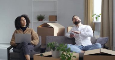 Etrafı karton kutularla çevrili çok ırklı bir çift kanepeye oturmuş yeni bir ev seçiyor dizüstü bilgisayar ve telefon planlaması ile ev arıyorlar mobilya paketlerini internetten sipariş ediyorlar.