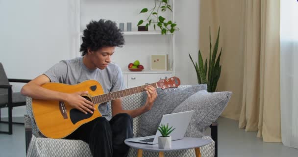 Дома учится играть на музыкальном инструменте афроамериканский парень-подросток студент-музыкант, сидящий на диване в гостиной и смотрящий на ноутбук онлайн-урок от наставника, играющего на гитаре создает музыку — стоковое видео