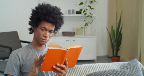 Afro american attractive focused smart student black guy sitting at home in modern living room reading orange book teaches subject enjoy leisure, education and knowledge concept, hobby and lifestyle — Stock Video