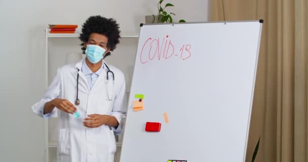 Genç, profesyonel afro-amerikan terapist, tıbbi maske takıyor. Üzerinde enfeksiyon virüsü hakkında yazılanlar var. Virüs, antibakteriyel jel dezenfektanının nasıl kullanılacağını gösteriyor. — Stok video