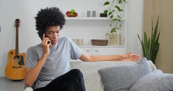 Serious confident Afro-American student teen guy talking on mobile phone indoors at home discussing news with friends answer answers question negatively solves problems sitting on sofa in living room — Stock Video