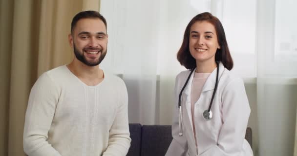 Dwoje ludzi kaukaska kobieta brunetka lekarz lekarz lekarz pracownik medyczny i tysiąclecia mężczyzna pacjent siedzi razem w klinice szpitalnej patrząc w aparacie stwarzając uśmiechnięty szczery, koncepcja odzyskiwania — Wideo stockowe
