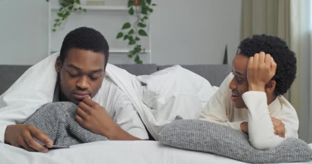 Père occupé se trouve avec sa femme sous les couvertures en regardant le téléphone mobile, fille soudainement drôle apparaît sous couverture enfant petite fille joue cacher et chercher avec ses parents dans le lit rit avec la mère — Video