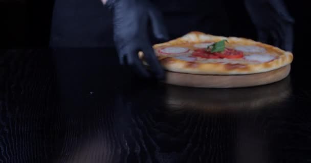 Gehakt shot van mannelijke handen van ober chef-kok in zwart uniform en latex handschoenen met houten ronde boord met hete verse lekkere kaas vegetarische pizza zet schotel op tafel te draaien, fast food concept — Stockvideo