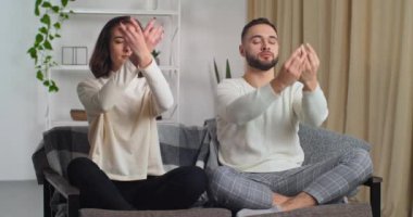 Mutlu çift evlerinde kanepenin üzerinde modern oturma odasında sabah antrenmanı yapıyor. Beyaz kadın ve erkek aynı hareketi yapıyor. Kolları nilüfer çiçeğinde oturuyor. Meditasyon yapıyor, ruhani dersler veriyor.