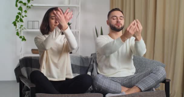 Gelukkig stel dat 's ochtends thuis oefent, moderne woonkamer op de bank. Blanke vrouw en man bewegende handen maken dezelfde beweging met armen zittend in lotushouding mediteert, spirituele oefeningen lessen — Stockvideo