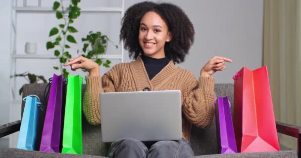 Счастливая улыбающаяся африканская женщина с ноутбуком сидит на диване, делая онлайн покупки онлайн заказы товары модная одежда обнимает яркие яркие сумки покупки радует успешное инвестирование — стоковое видео