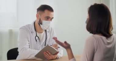 Kadın hasta terapiste baş ağrısı, sırt ağrısı, semptomlar hakkında şikayette bulunuyor. Doktor muayenesinde oturan, yüz maskesi takan ve deftere yazan uzman bir erkek doktor.